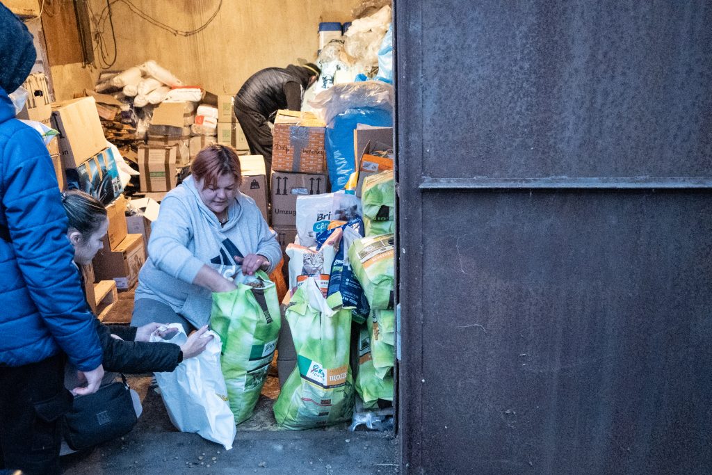 Woman kneeling by large dog food bags, seen through the open door of a shed