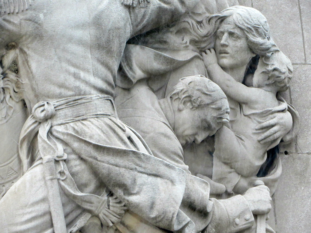 White (perhaps marble) statue showing a man, woman, and child cowering as another figure marches past.