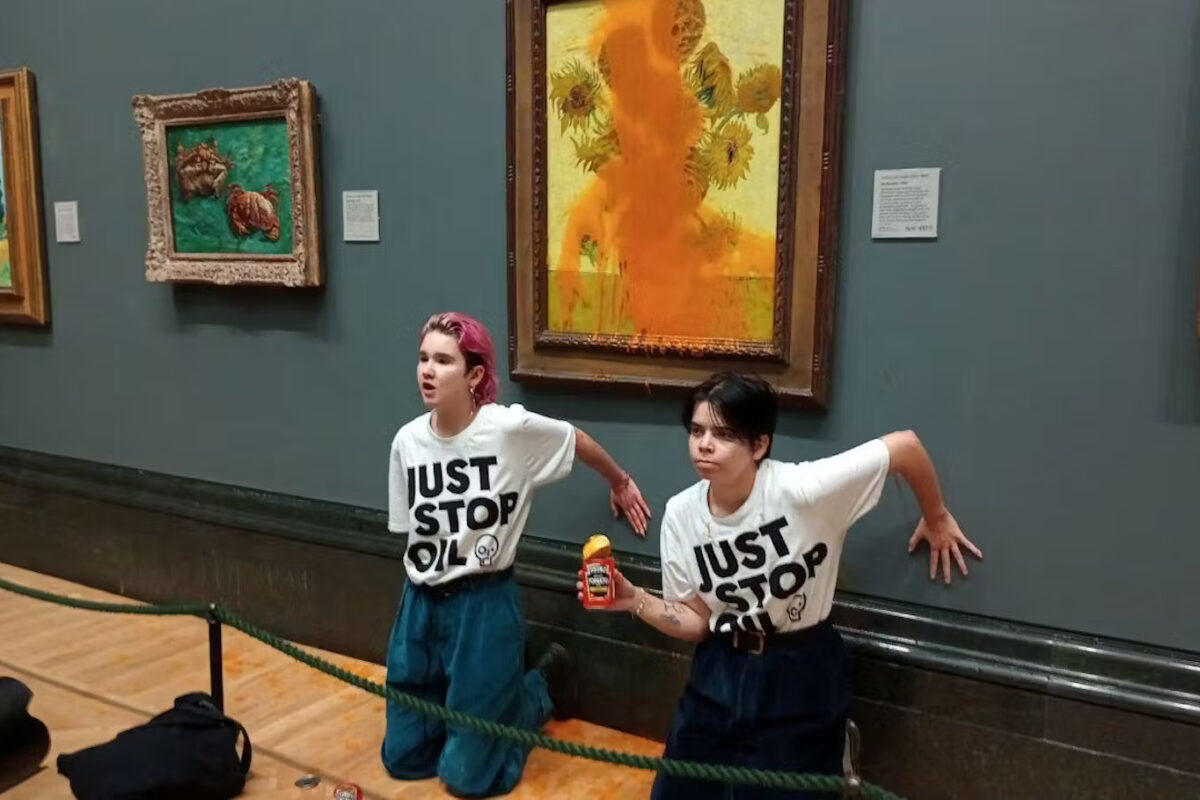 Two Just Stop Oil protestors kneel in front of the Van Gogh sunflowers painting that they have just covered in soup. One holds the empty can of soup in their hand.