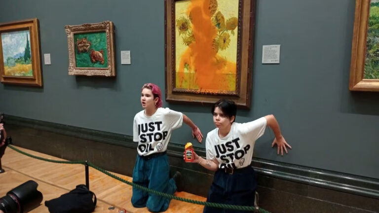 Two Just Stop Oil protestors kneel in front of the Van Gogh sunflowers painting that they have just covered in soup. One holds the empty can of soup in their hand.