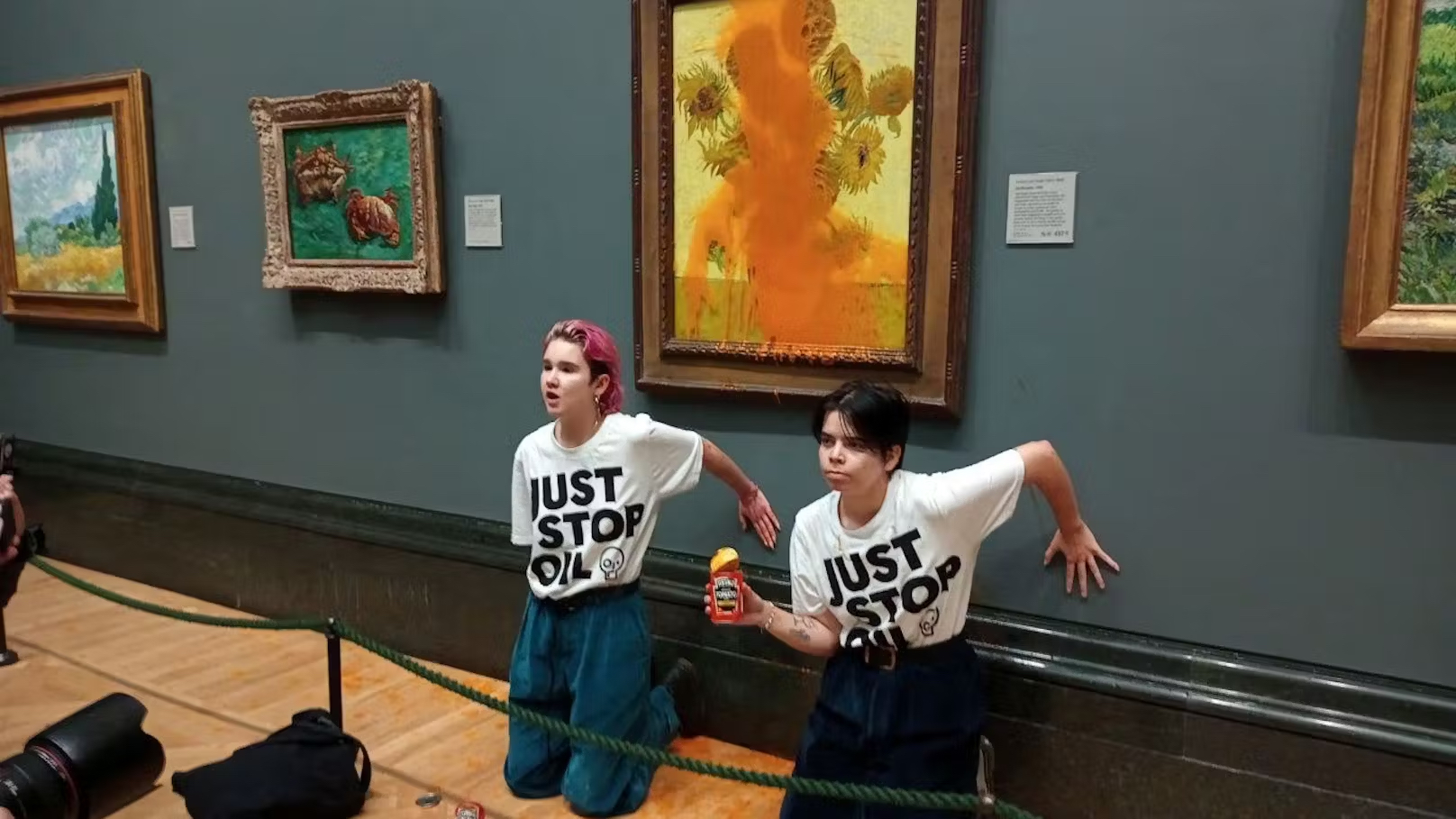 Two Just Stop Oil protestors kneel in front of the Van Gogh sunflowers painting that they have just covered in soup. One holds the empty can of soup in their hand.