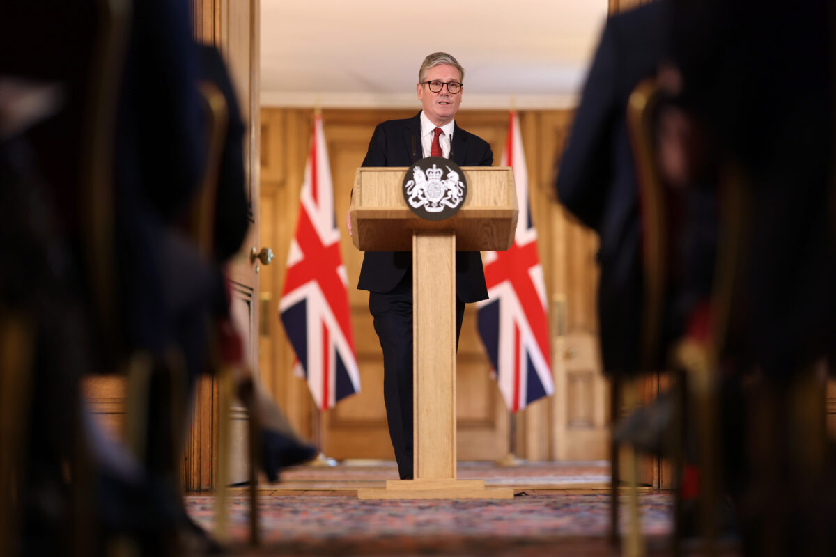 UK Prime Minister Keir Starmer stands at a wooden podium with the official government crest, addressing the media during a press conference. Behind him are three Union Jack flags and wooden paneling, while blurred figures of seated attendees are visible in the foreground.