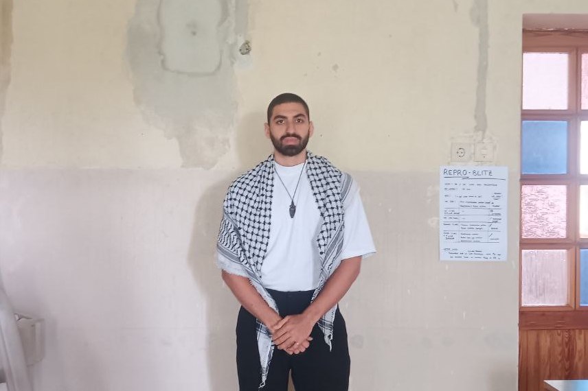 Yasan Eissa stands in front of a wall, hands clasped in front of him. He is wearing a keffiyeh and a necklace with a pendant.