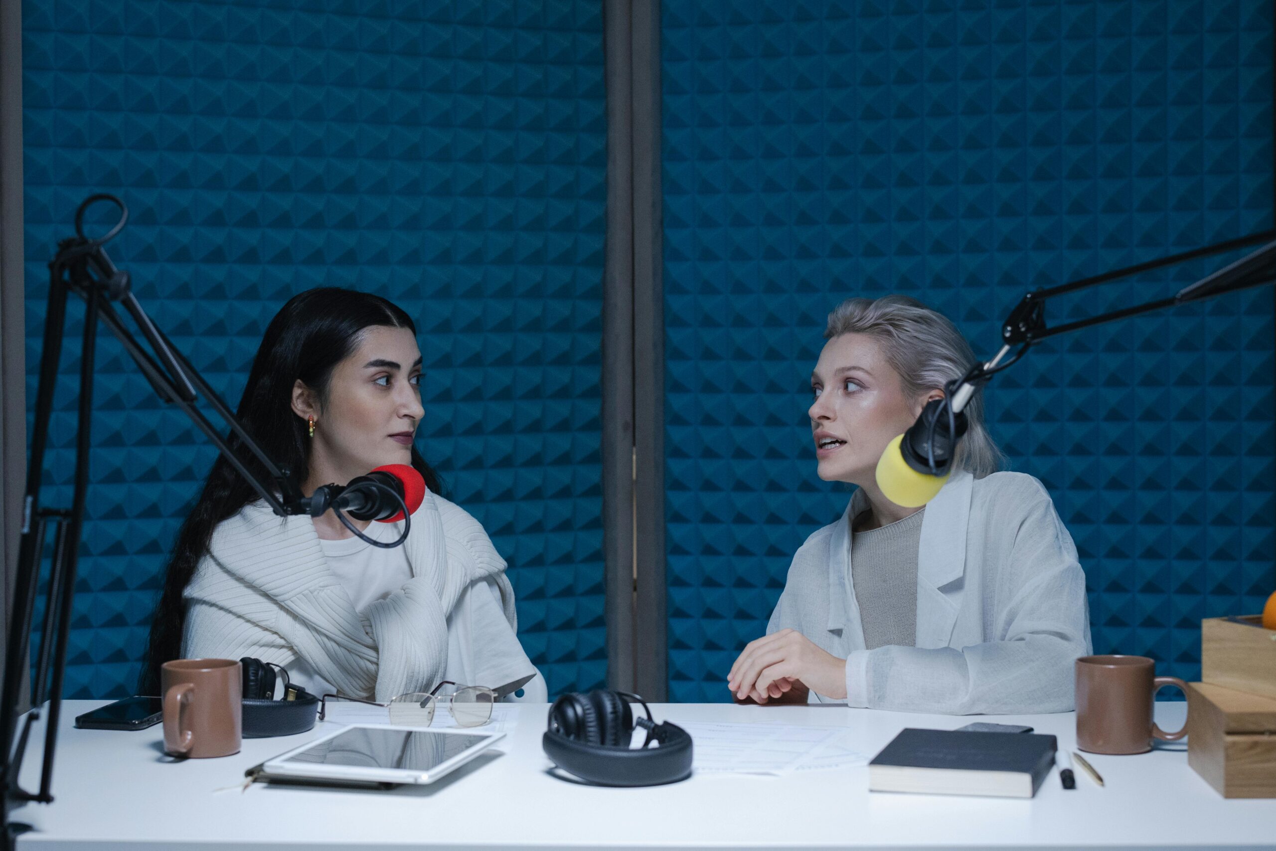 Two people sit in a podcast setting with mics. One is listening and the other is speaking with intensity.