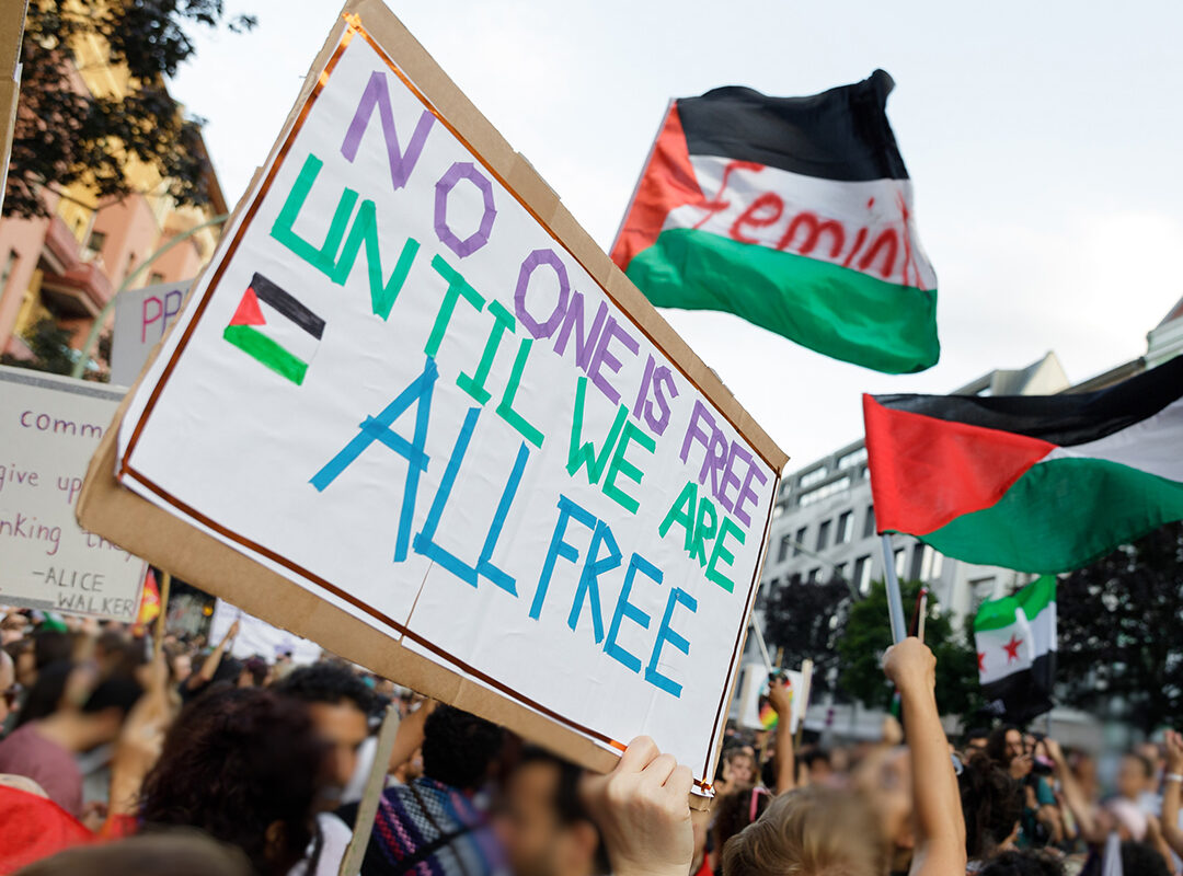 Hands in a crowd hold up Palestinian flags and a sign saying "No one is free until we are all free."