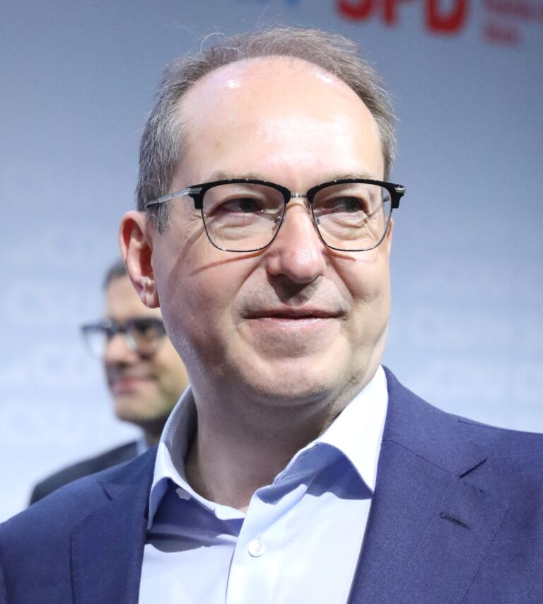 Alexander Dobrindt wearing glasses and a blue suit, standing in front of a blurred background with the logos of CDU and SPD partially visible.