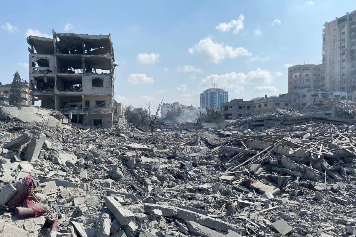 Widespread destruction in Gaza following Israeli airstrikes, showing a collapsed urban area with piles of rubble, twisted metal, and remnants of buildings. A partially standing, severely damaged multi-story building is visible to the left, surrounded by debris and dust, while modern high-rises appear intact in the distant background under a blue sky.