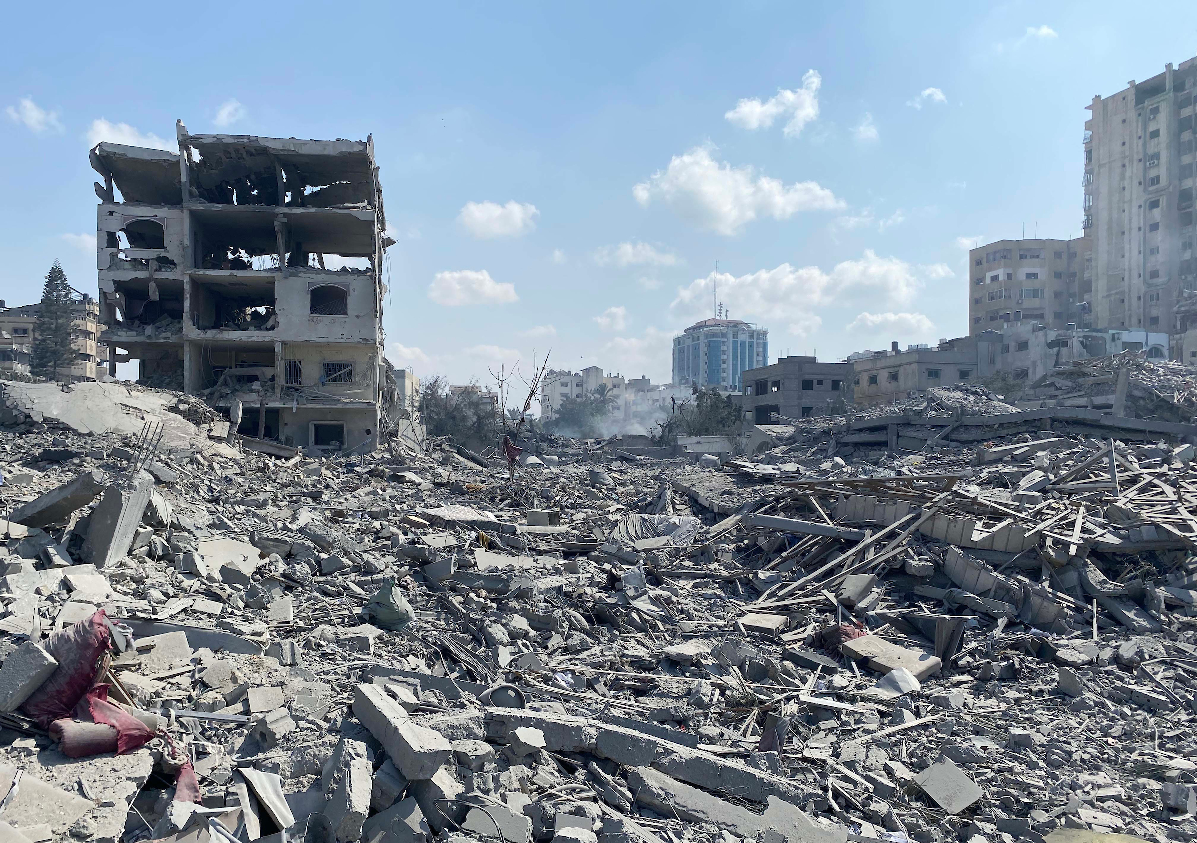 Widespread destruction in Gaza following Israeli airstrikes, showing a collapsed urban area with piles of rubble, twisted metal, and remnants of buildings. A partially standing, severely damaged multi-story building is visible to the left, surrounded by debris and dust, while modern high-rises appear intact in the distant background under a blue sky.
