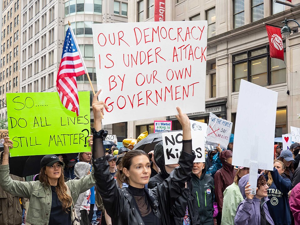 A photograph of protestors at a "No Kings" demonstration. One holds a sign which reads "Our democracy is under attack by our own government".