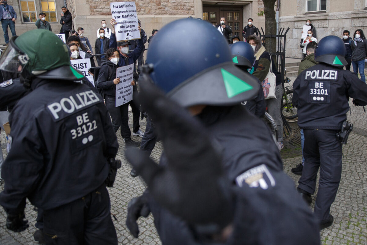 A group of police with helmets and heavy-duty jackets stand between the camera and a group of protestors. The closes police holds their hand in front of the camera, as if to say "don't film me"