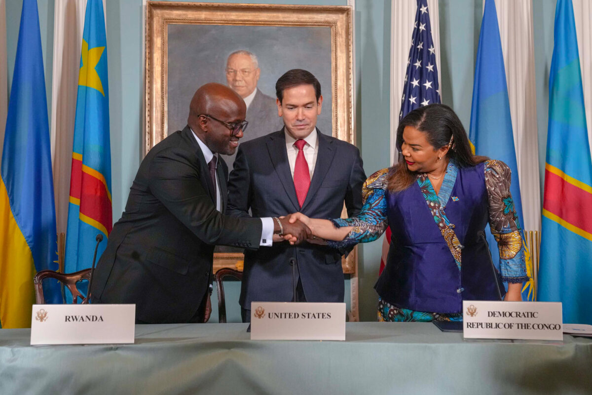 Rwandan and Congolese delegates shake hands across a table as the U.S. representative looks on, with national flags and a portrait in the background.