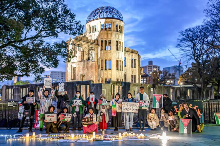 A group of 17 people stand in front of the Hiroshima Atomic Bomb memorial holding various signs. There are candles and signs on the ground as well.