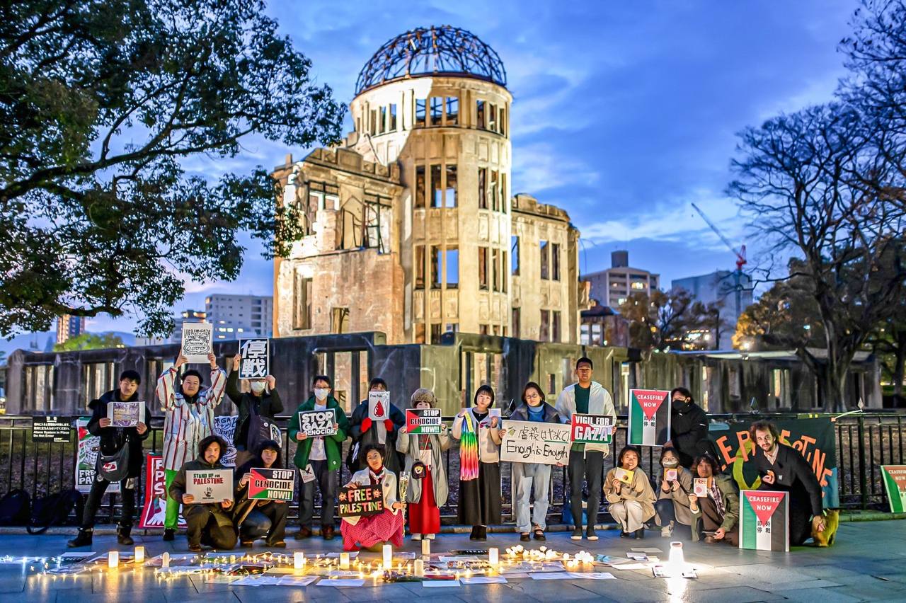 A group of 17 people stand in front of the Hiroshima Atomic Bomb memorial holding various signs. There are candles and signs on the ground as well.