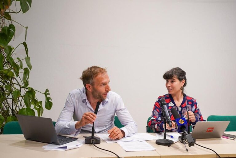Two people sit at a press conference, with computers and microphones.
