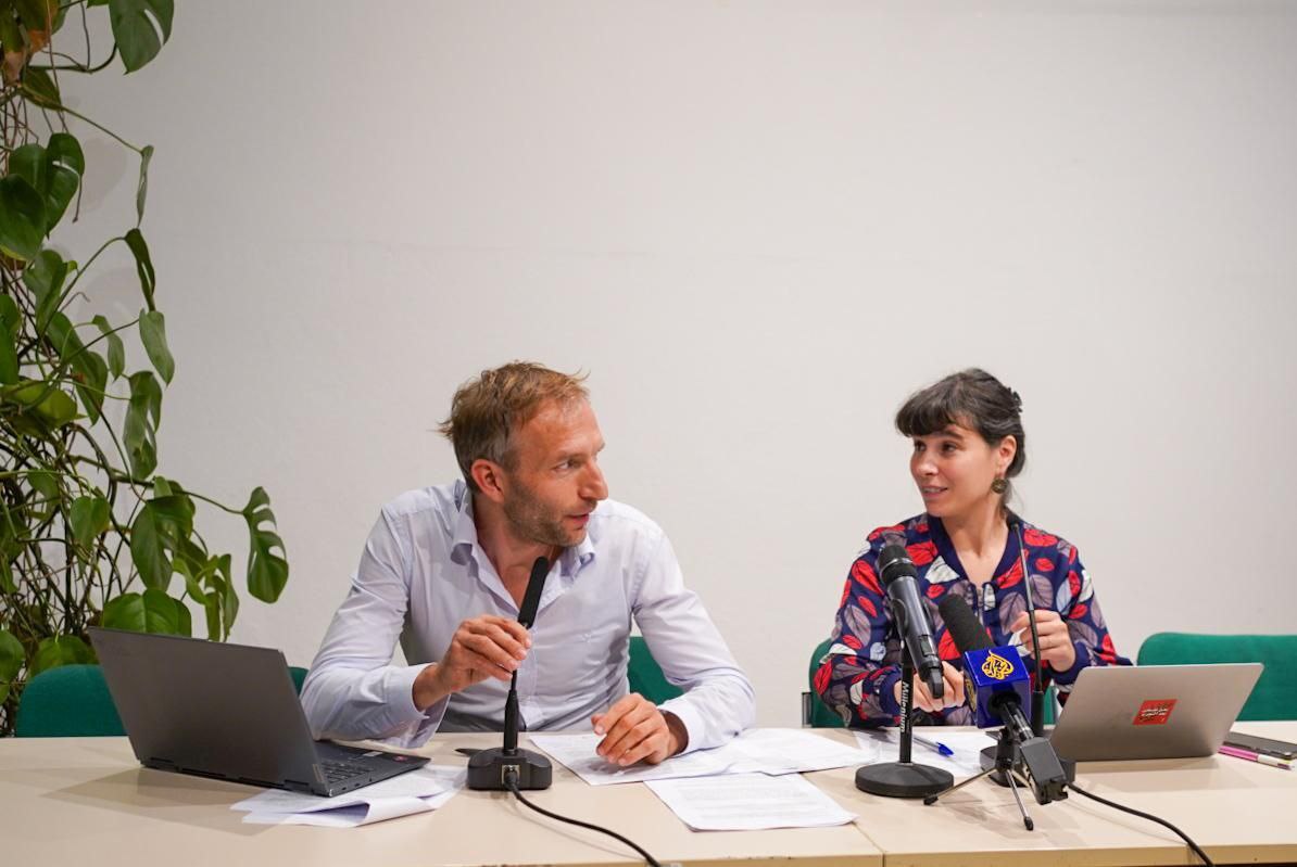 Two people sit at a press conference, with computers and microphones.