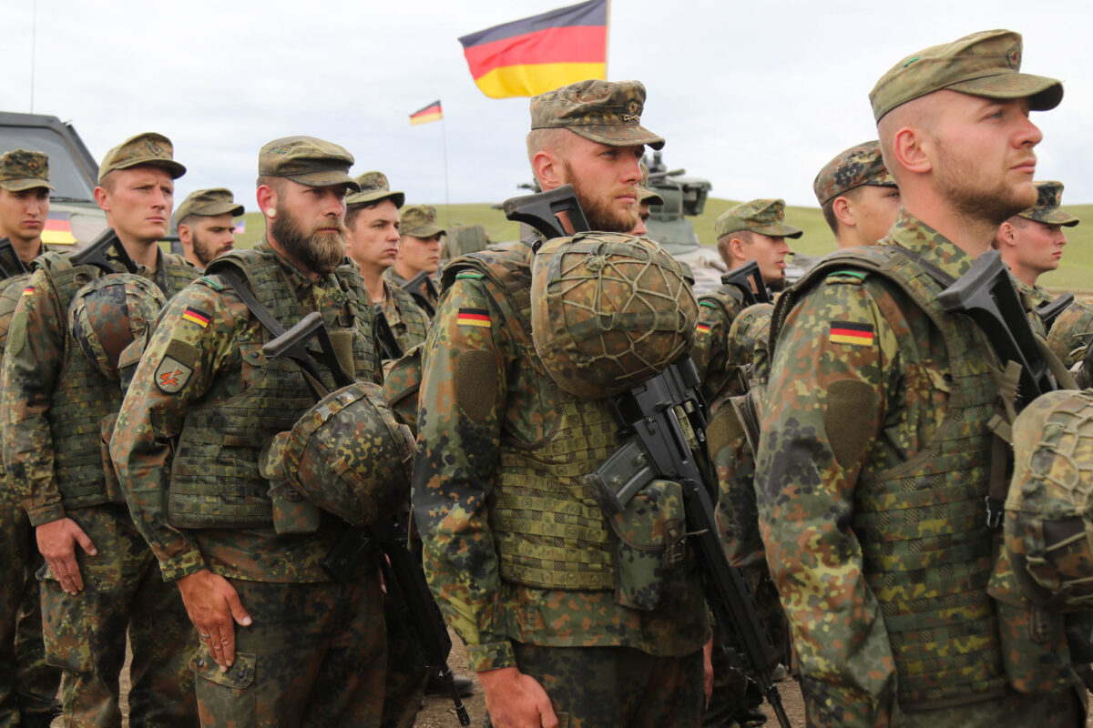 A crowd of soldiers stand in formation. A German flag flies overhead.