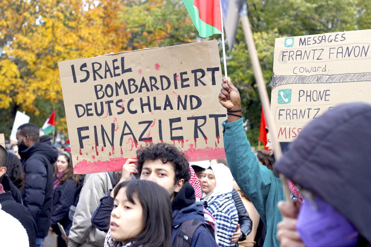 People at a protest, waving Palestine flags in the background. One person wearing a Keffiyeh, holds a sign that reads: Israel bombardiert, Deutschland finanziert.