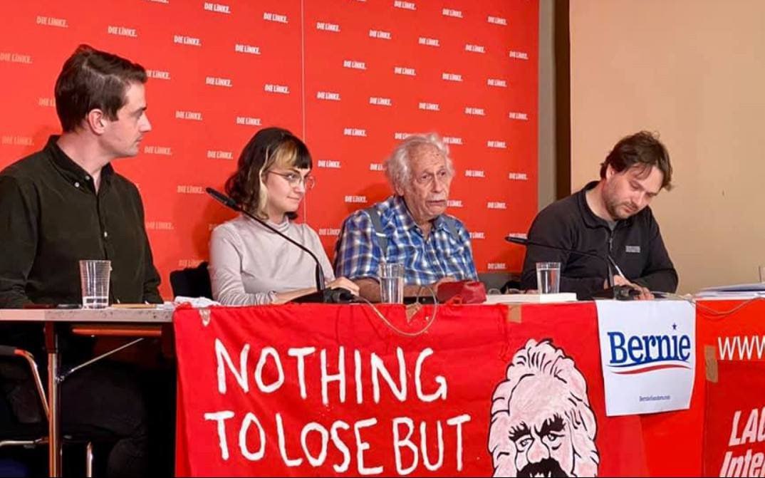 Victor Grossman sits a table with 3 other panelists. The banner hanging from the table is red with white lettering that says "Nothing to lose but" and the rest is cut off. The banner also shows part of Karl Marx's face and a Bernie campaign symbol.