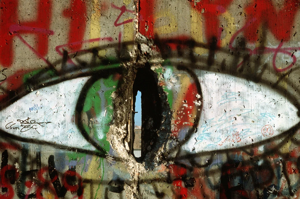 Graffiti eye with red and green iris. There is a huge gash cutting right through the eye and showing view of something on the other side of the wall.