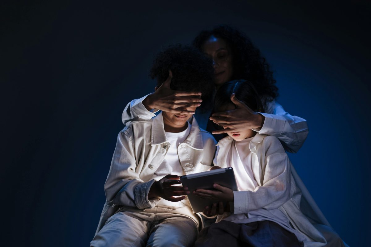 Adult covering the eyes of two children who are holding a tablet.