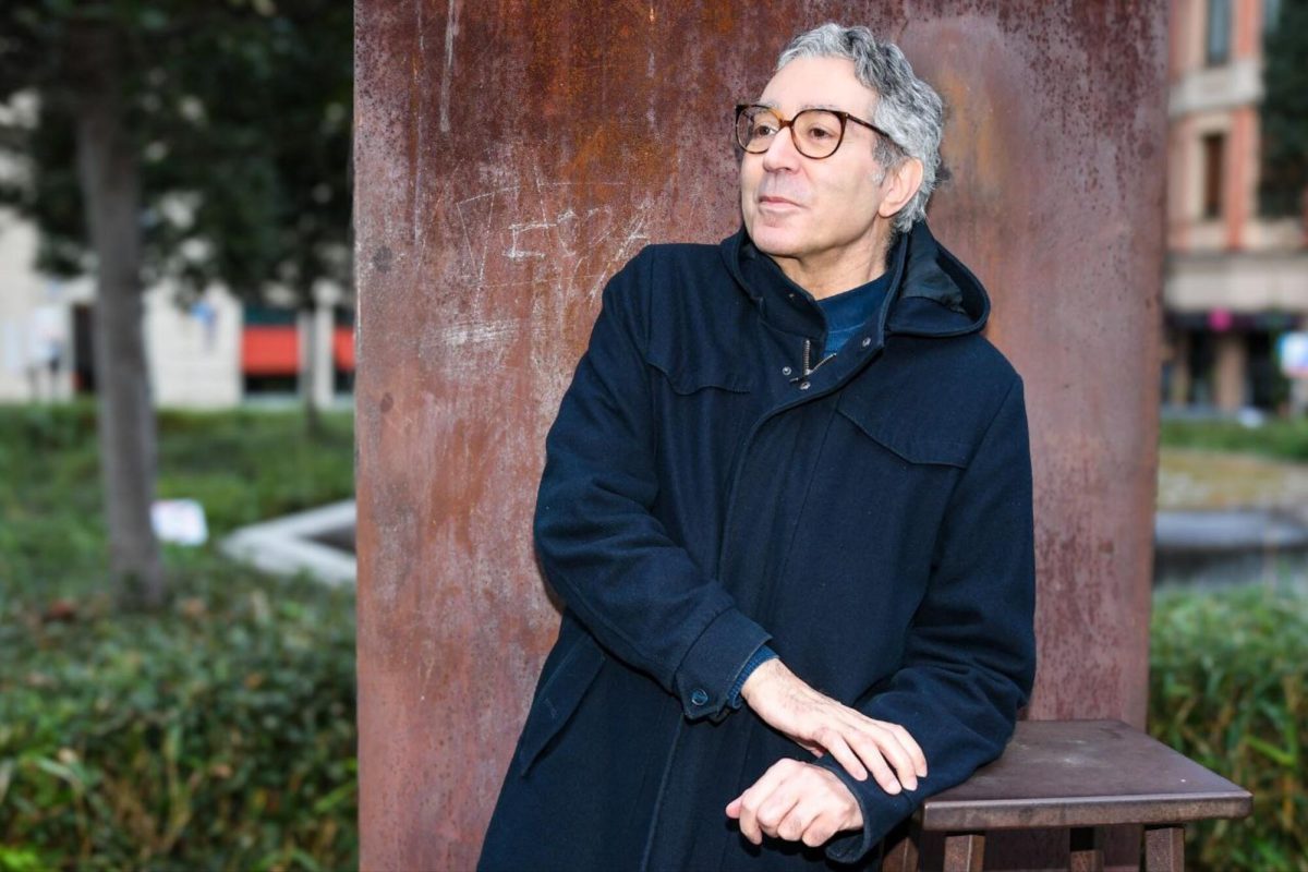 Didier Eribon leans one arm on a small table. He is wearing a dark coat, zipped almost all the way. He is looking off the the left, eyes shining and smiling slightly, looking as if he is about to say something. He is standing in front of a large rusted metal column at a park.