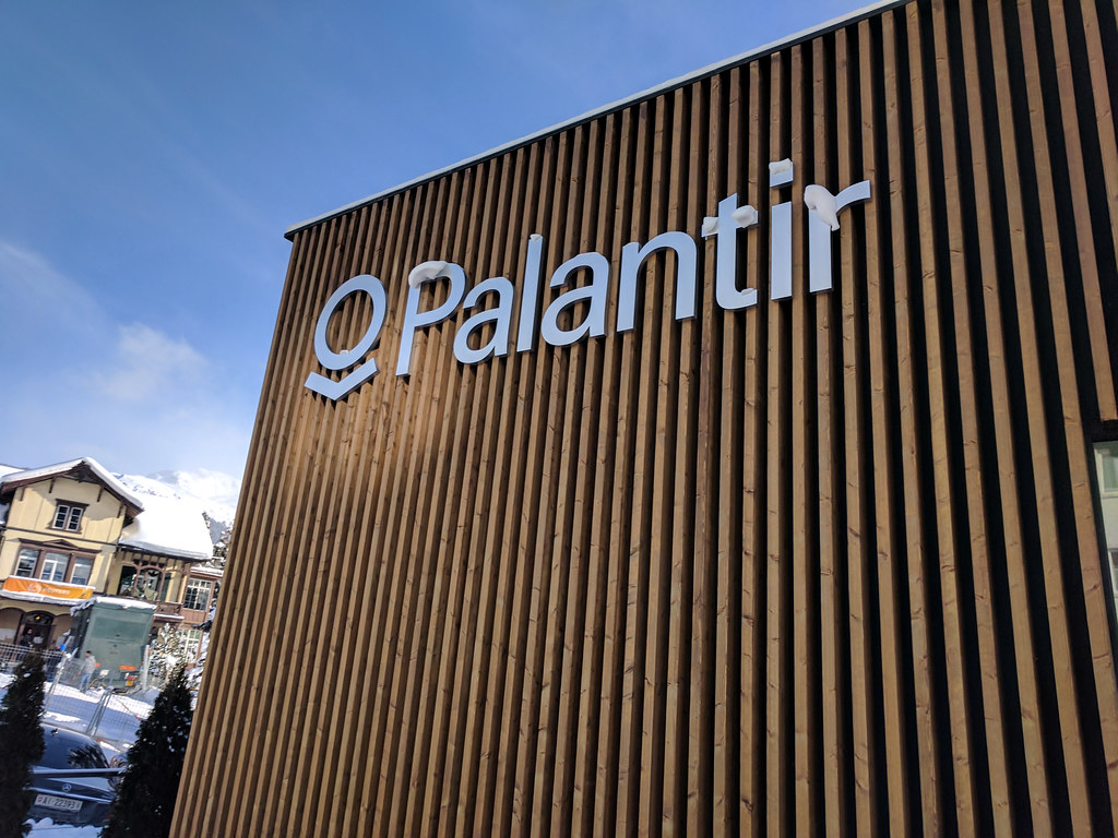 The side of a building with the Palantir logo and name in silver letters. There are bits of snow sitting on some of the letters, and some houses with snowy roofs in the background.