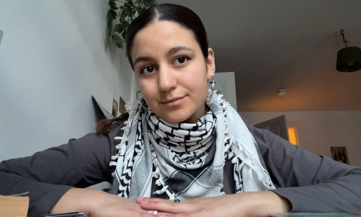 Dima is seated with arms resting on a table. She is looking right at the camera but her face is tilted slightly to the left. Her mouth curves into a slight smile. She wears a kuffiyeh around her neck.