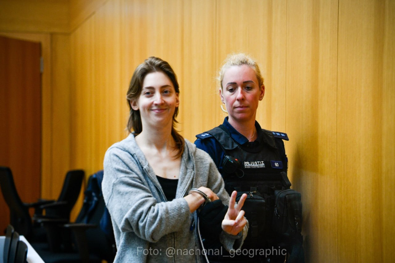 Vi Kovarbasic holding up a peace sign in court, accompanied by an officer.