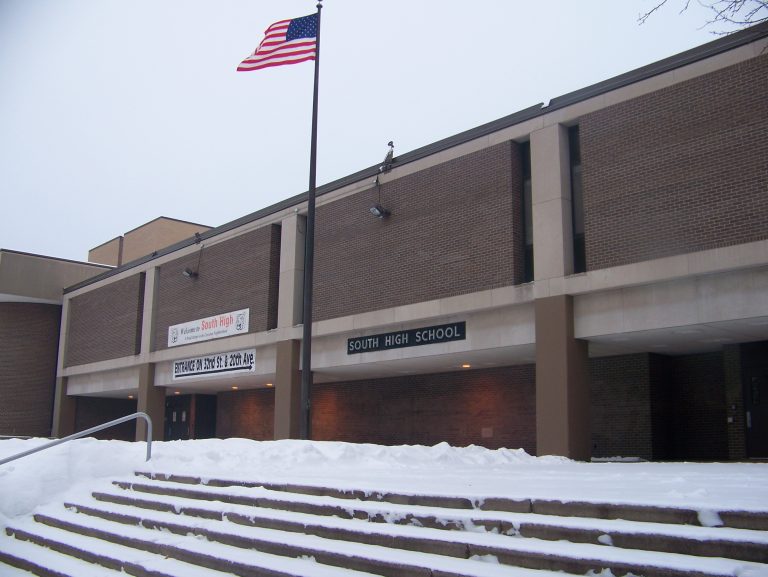 The entrance of Minneapolis South High School. There is a US flag flying right above. The steps leading up to the entrance are covered in snow.