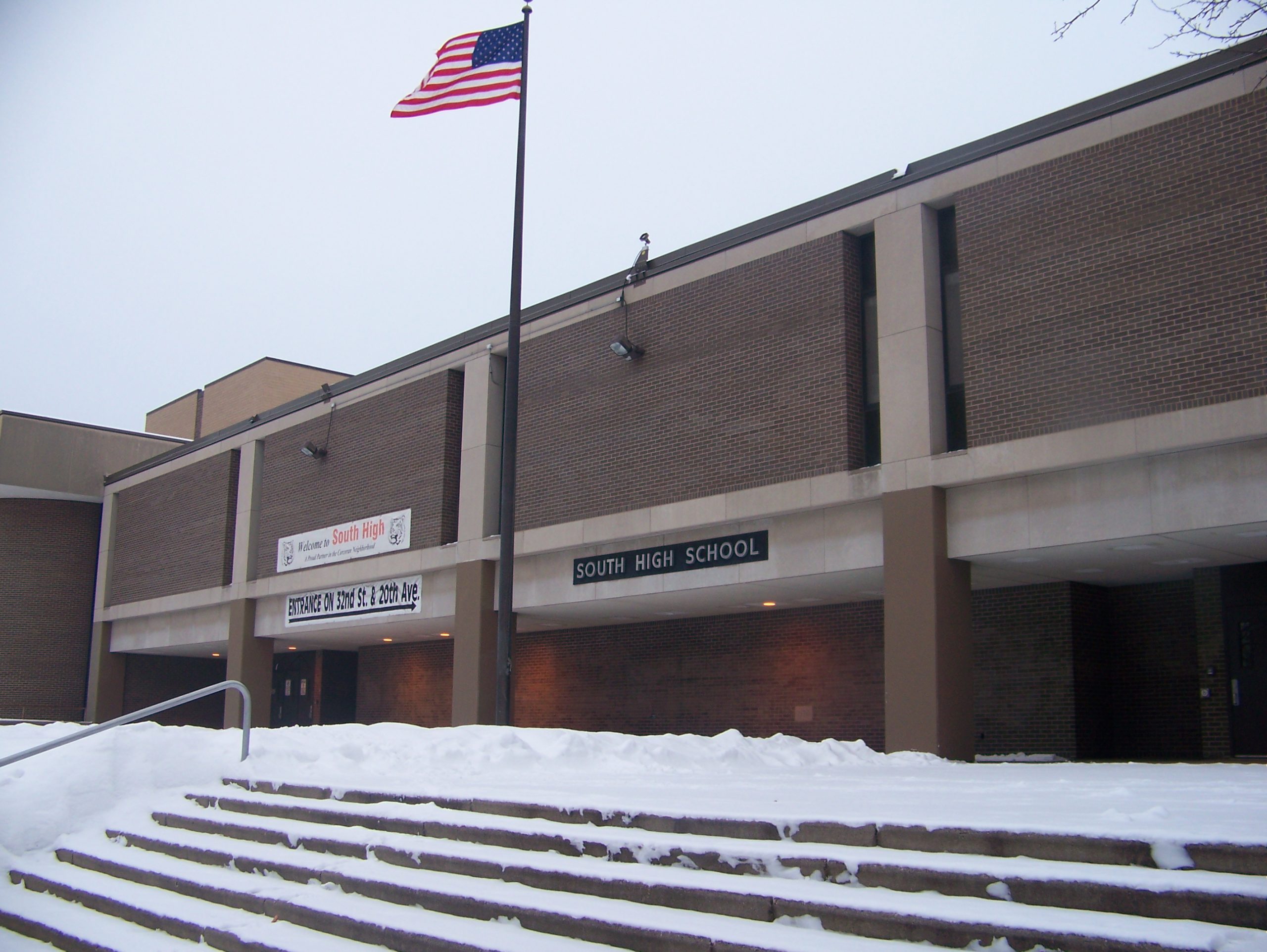 The entrance of Minneapolis South High School. There is a US flag flying right above. The steps leading up to the entrance are covered in snow.