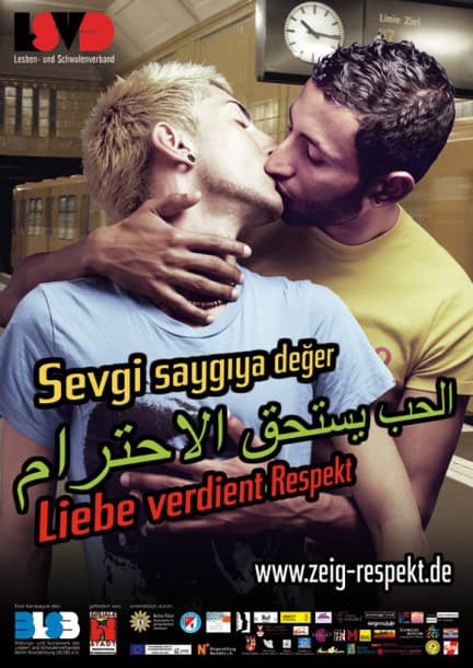 Two men are engaged in a passionate kiss. The man in front is blond and wears a light blue T-shirt. The man behind behind him is a brunette, wearing a yellow shirt. The man behind has his right hand resting on the first man's neck. The phrase "Love deserves respect" is written in Turkish, Arabic, and German.