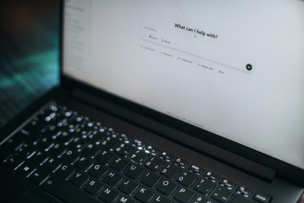A laptop open to a white chatbot screen. The words "What can I help with?" with an empty prompt input below.