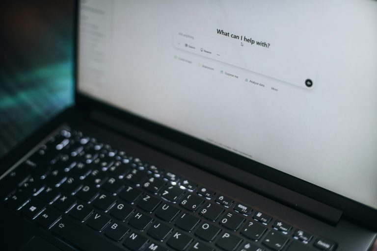 A laptop open to a white chatbot screen. The words "What can I help with?" with an empty prompt input below.