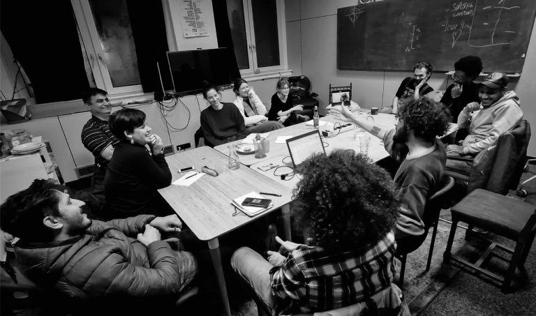 Black and white image of 11 people sit around a table, perhaps in a classroom since there is a chalkboard on one of the walls. Some are laughing, one is gesticulating. There are notebooks and laptops on the table.