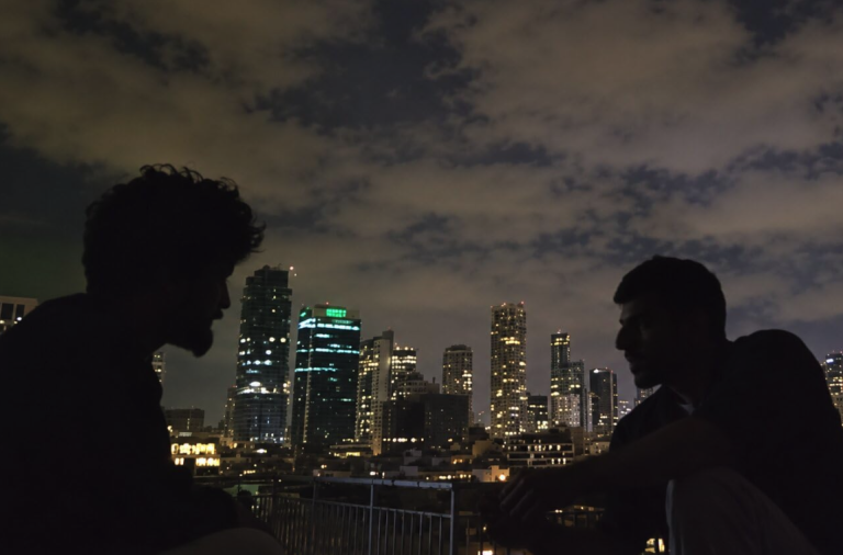 Two silhouettes of men against a cloudy night sky and a city scape. The men appear to be sitting and facing each other, perhaps talking. It is a still from the trailer of the movie.