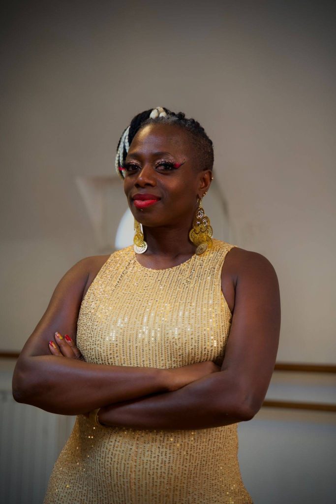 Maïmouna Coulibaly stands with arms crossed over her chest. She is looking at the camera with confidence and a subtle smile. Her braided hair is gathered up behind her head although a few light blue braids are visible. She has pink glittery eyeshadow, a hint of red in her eyelashes, and bright red lipstick. She is wearing a yellow sequin dress.